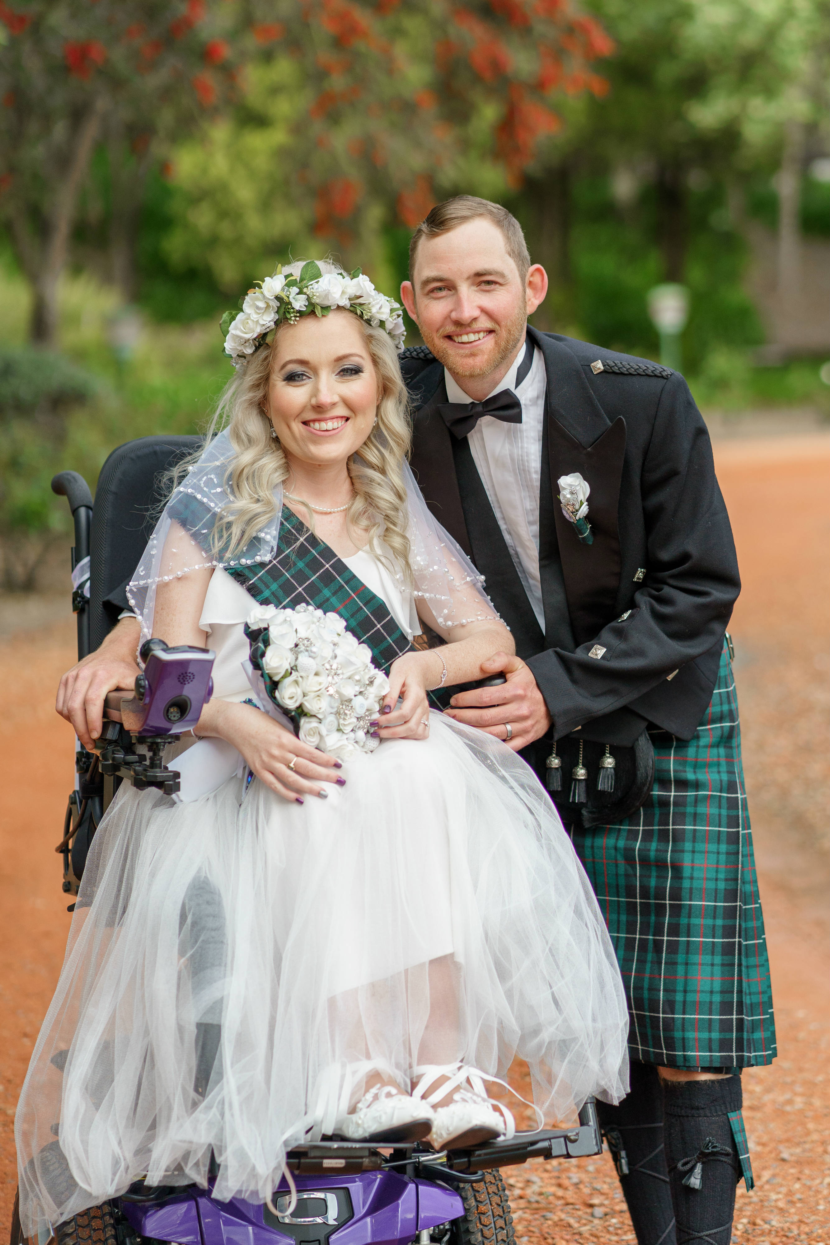 wedding day photo of couple, the young woman is in an ivory wedding dress, green tartan sash and in a purple wheelchair, her husband has a black jacket, bowtie and green kilt.