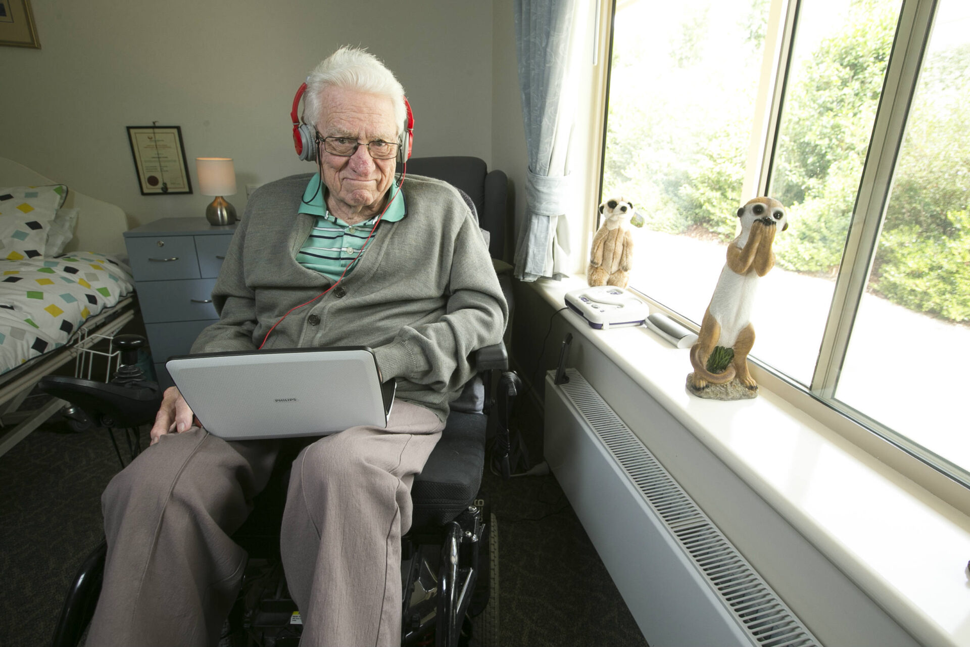 This is a photo of Gordon, in his room, sitting in a wheelchair next to his bed and beside a window. Gordon is wearing headphones and has a laptop on his lap. He looks into the camera with a wry smile. On the window sill are one stuffed toy meercat and one statue of a meercat.