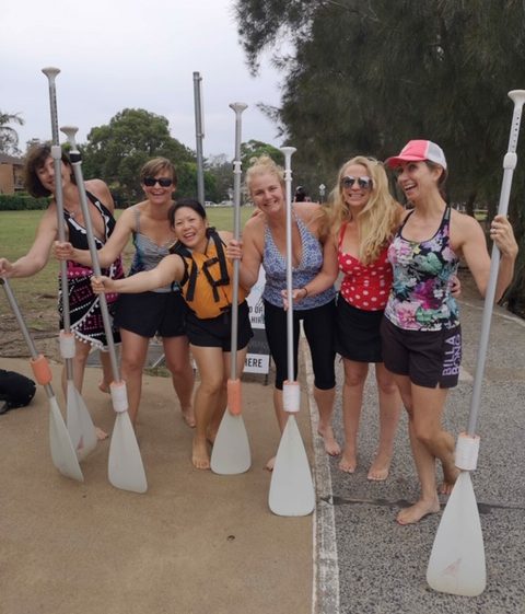 6 women posing, smiling, each holding a paddle. They are dressed in swimwear and shorts.