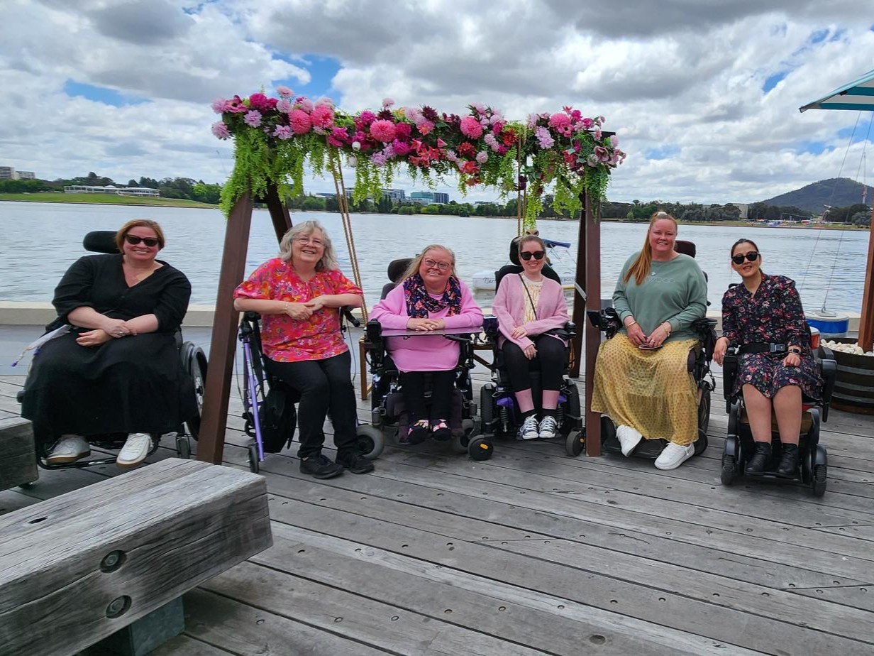 A group of people sit together on a wooden deck by a lake under a cloudy sky. A floral arch decorates the background, creating a joyful atmosphere.