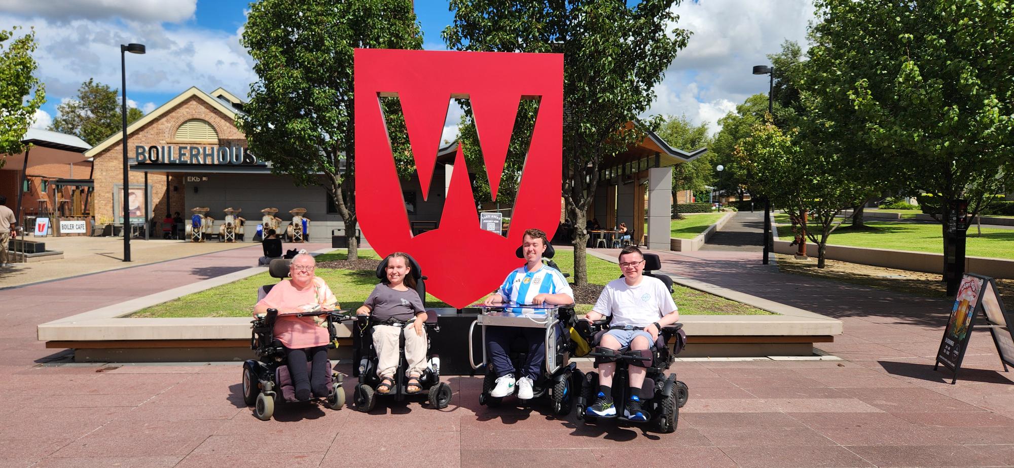 Four people in wheelchairs smile in front of a large red "W" sculpture outdoors. The background features trees and a building labeled "Boilerhouse."
