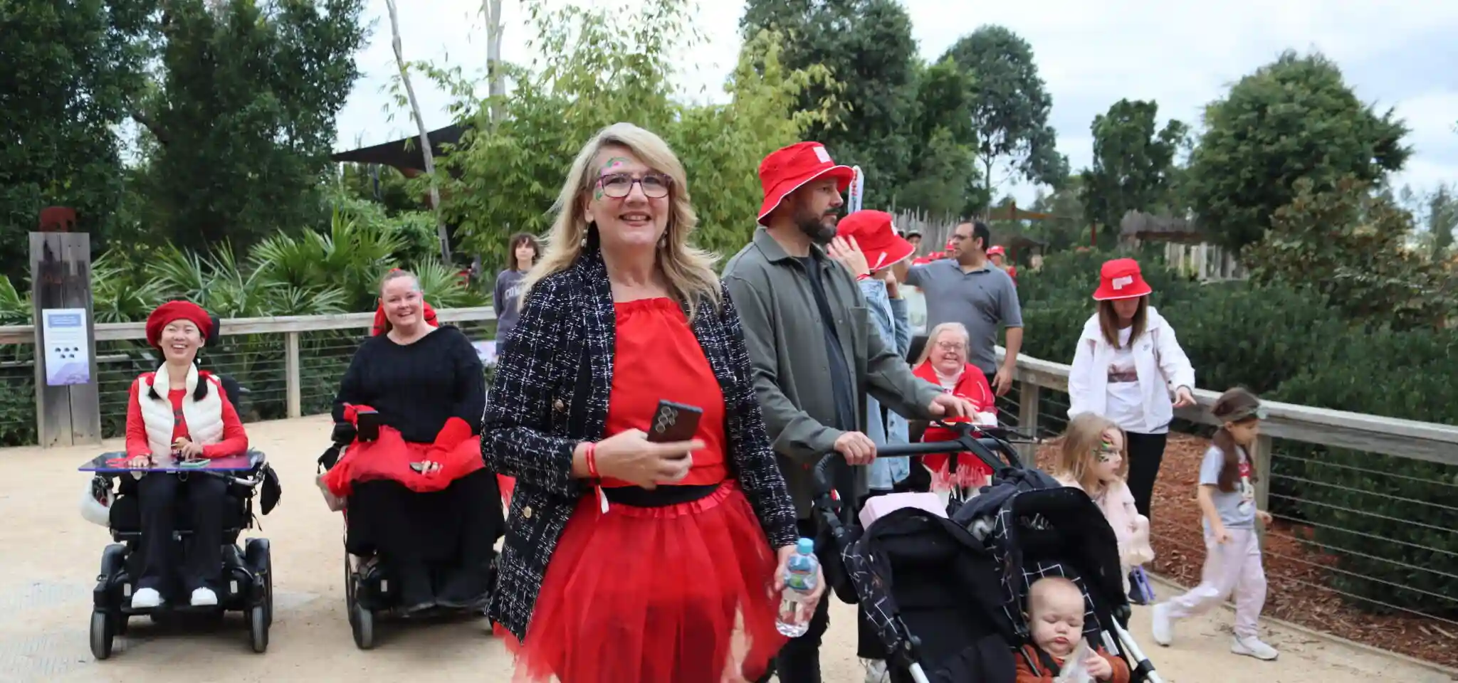 A group of people, including adults, children, and individuals using wheelchairs, walk outdoors on a dirt path wearing red clothing and accessories during Muscular Dystrophy NSW's Big Red Roll & Stroll 2025 event. Trees and greenery of Sydney Zoo line the background.