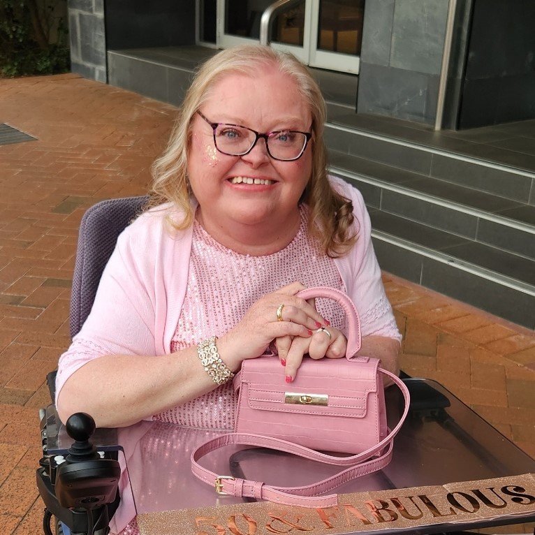 Woman in pink sparkly dress and cardigan, seated in a wheelchair, smiles warmly. She holds a pink purse on her lap, conveying joy and elegance.