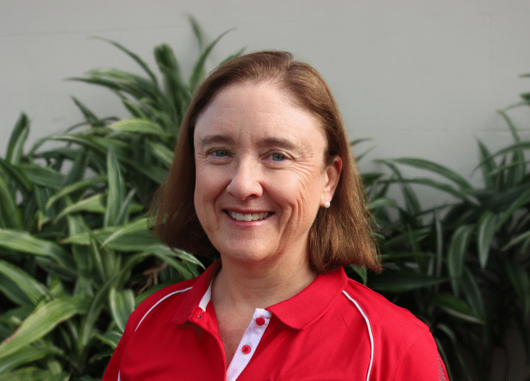A smiling woman with short brown hair wearing a red polo shirt stands in front of lush green plants, conveying a cheerful and welcoming mood.