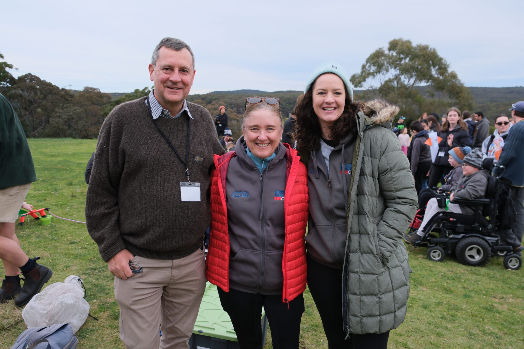 Three people, two women and one man, smile outdoors in warm jackets. A crowd and trees are in the background, suggesting a cheerful group event.