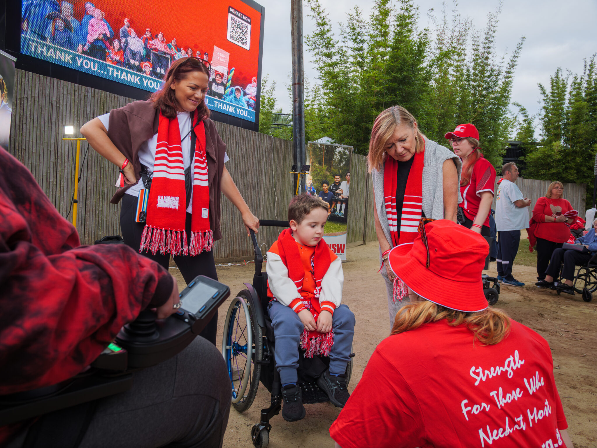 A smiling woman with a red scarf stands beside a boy in a wheelchair, wearing matching scarves. A seated woman in red talks to him. A large screen in the background shows a thank-you message. The scene conveys warmth and community support.