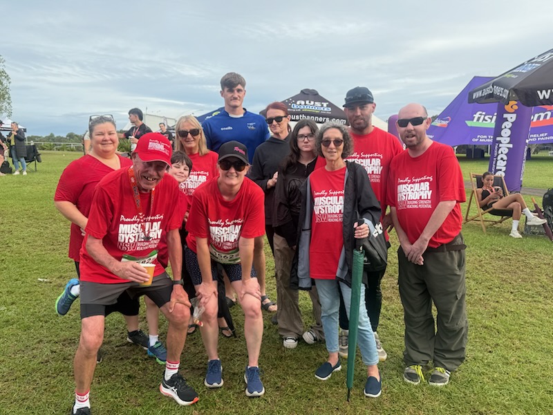 A group of people in red shirts stand on grass, smiling at a Muscular Dystrophy charity event. Tents and others in the background suggest a lively atmosphere.