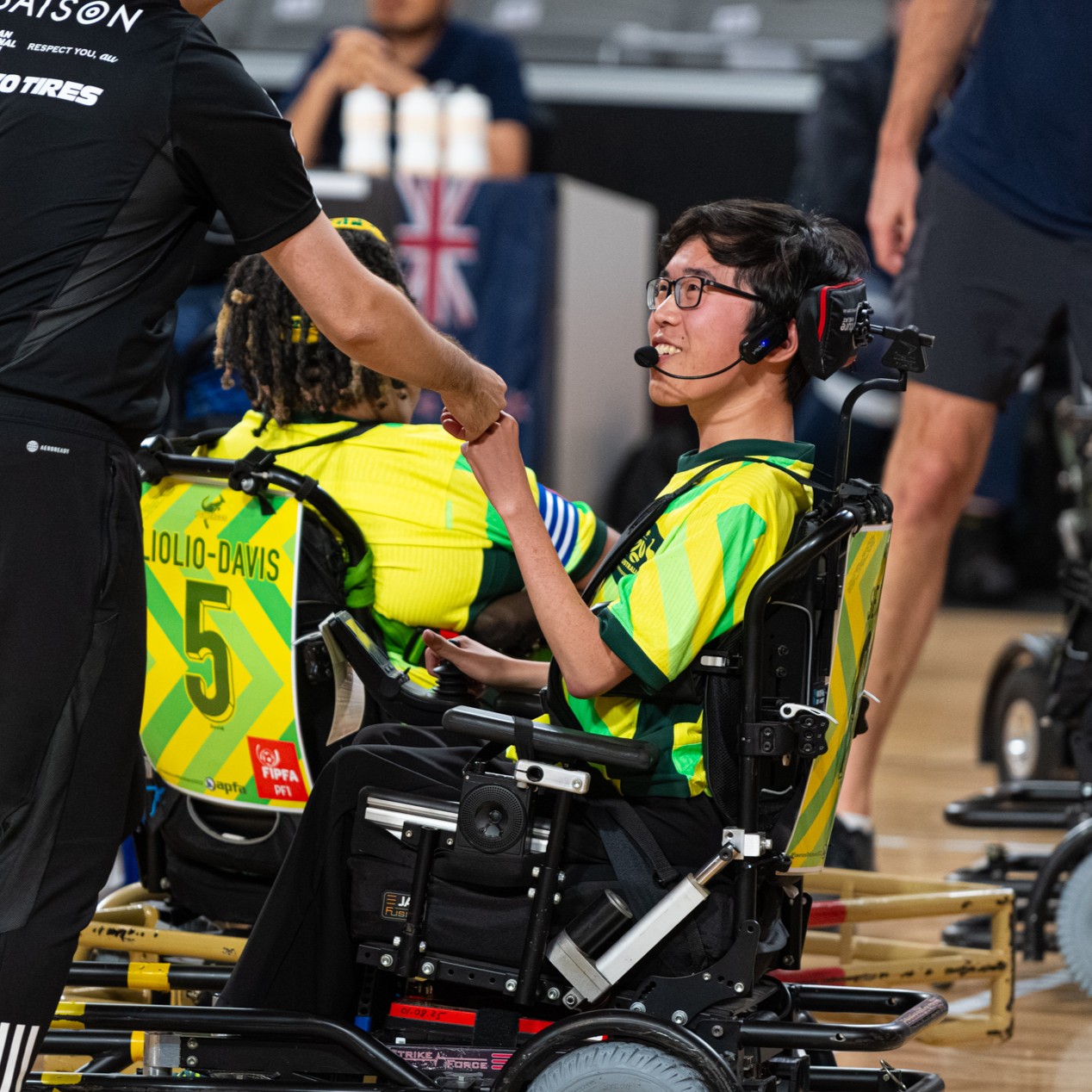 A young athlete in a yellow and green jersey smiles while fist bumping a coach. They sit in a power wheelchair during a sports event.