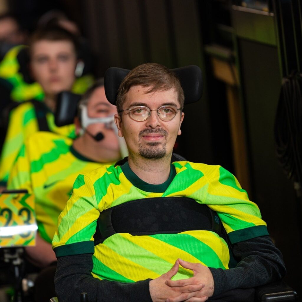 A man in a sports wheelchair, wearing a yellow and green jersey, smiles confidently. Teammates in the same outfit are visible in the background.