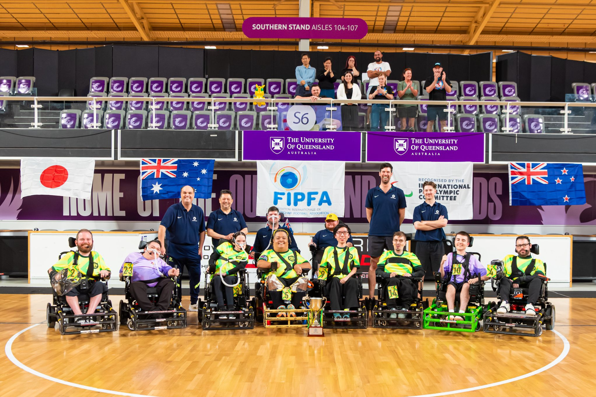 A wheelchair sports team poses on a court with smiling supporters behind. Banners and flags from Japan, Australia, and New Zealand are displayed.
