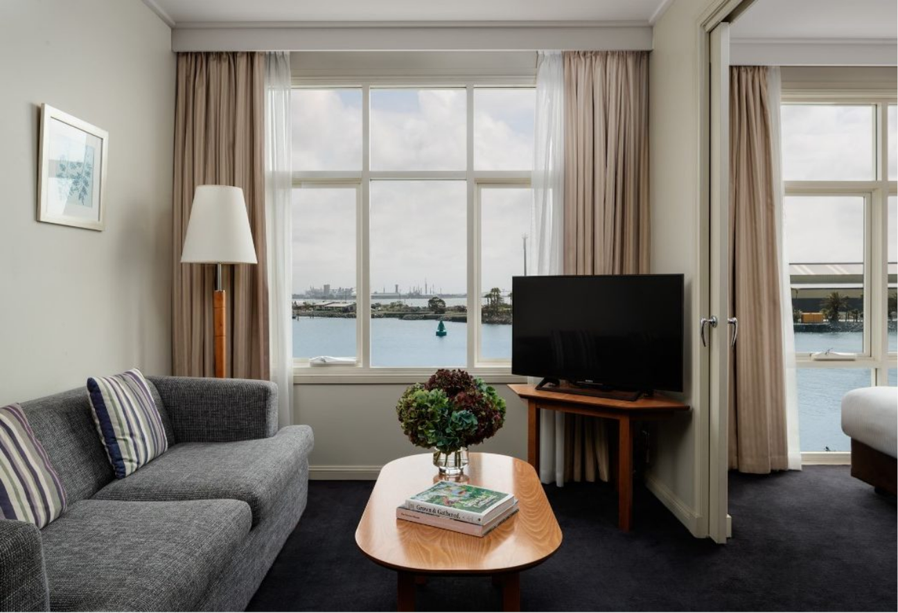 A cozy living room with a gray sofa and striped cushions faces large windows overlooking a serene waterfront view. A TV sits on a wooden stand, and a coffee table with a plant and book adds warmth.